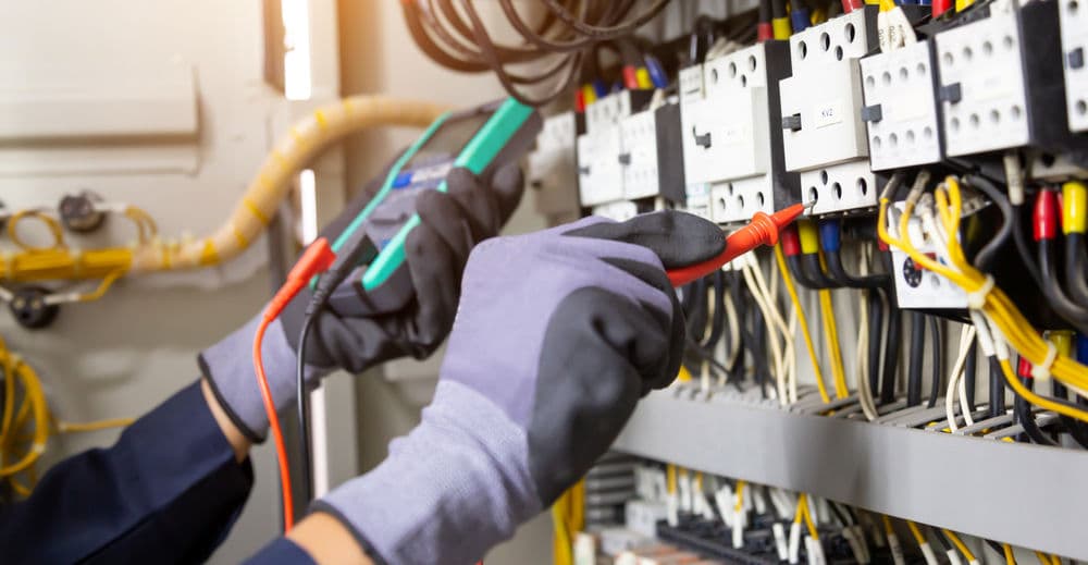 Electrician measuring voltage on circuit breakers with multimeter in electrical panel.